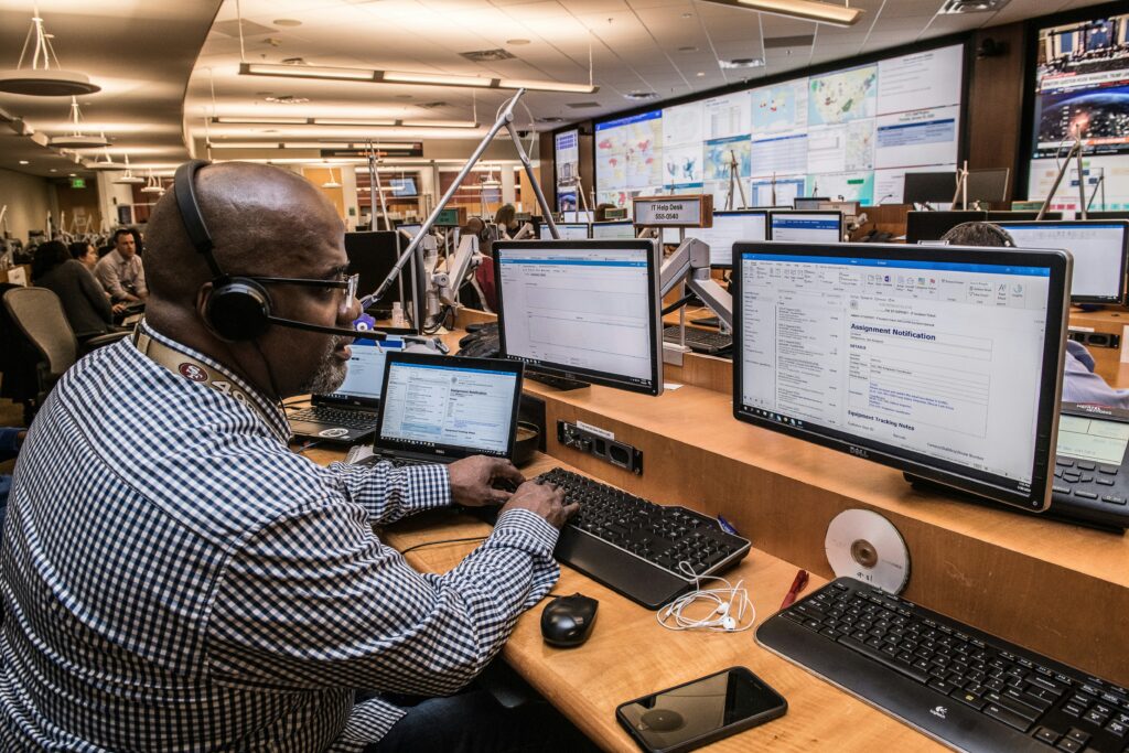 Man wearing a headset working at a multi-monitor workstation in a busy emergency operations center.
