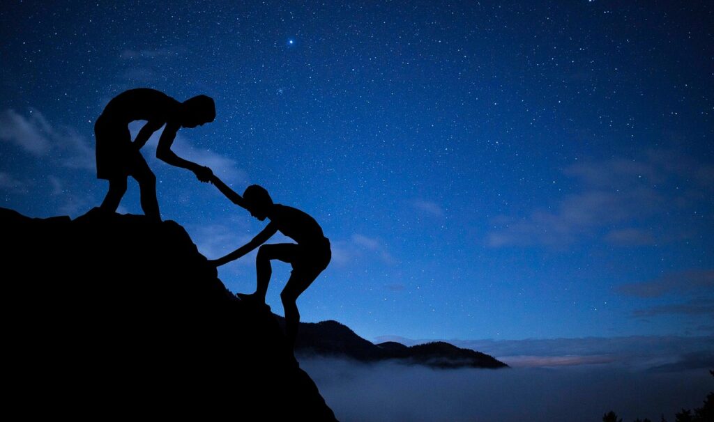 Silhouette of one person helping another climb a mountain under a starry night sky.