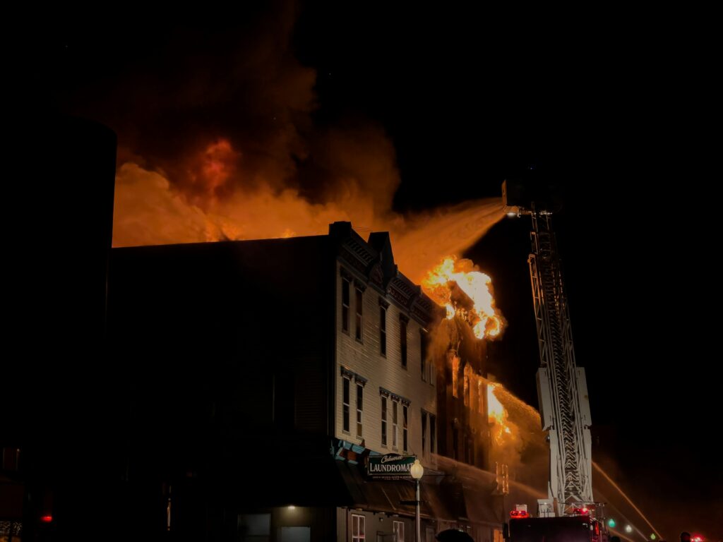Ladder truck sprays water on fully involved multi-story building fire at night.
