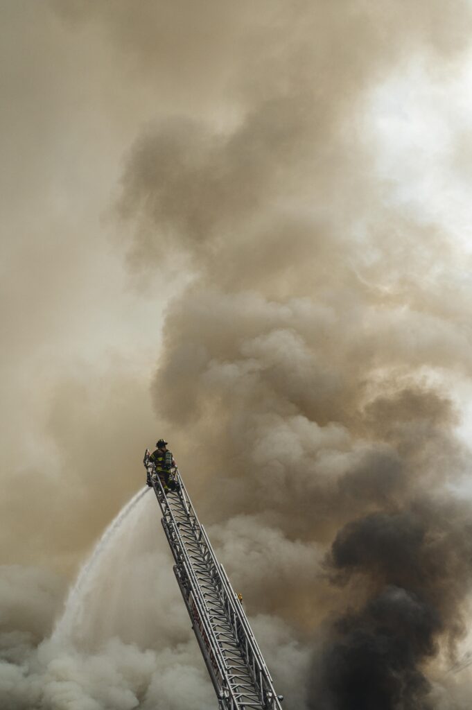 Firefighter atop an aerial ladder spraying water into thick smoke during a large-scale fire.