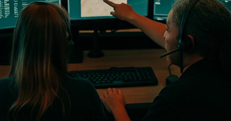 Experienced dispatcher providing one-on-one training to a new recruit while reviewing incident maps and CAD data on a triple-monitor workstation.