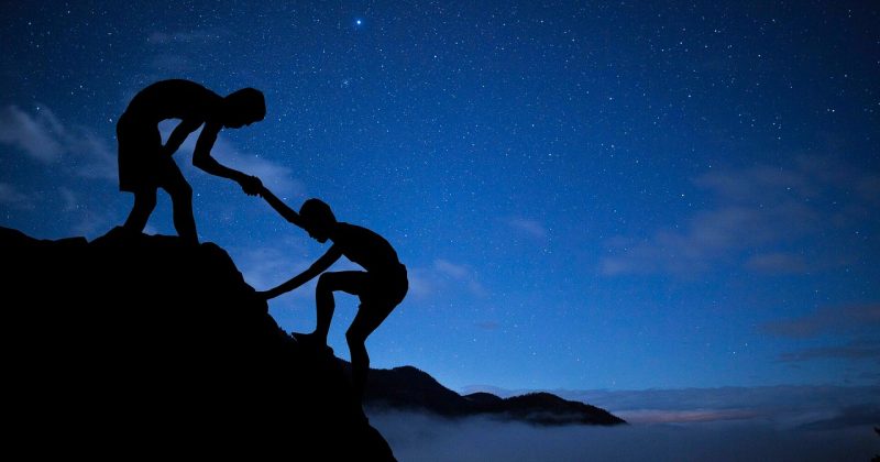 Silhouette of one person helping another climb a mountain under a starry night sky.