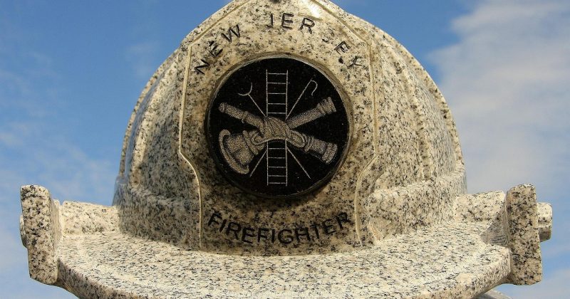 Granite firefighter helmet monument engraved with “New Jersey Firefighter” under a blue sky.