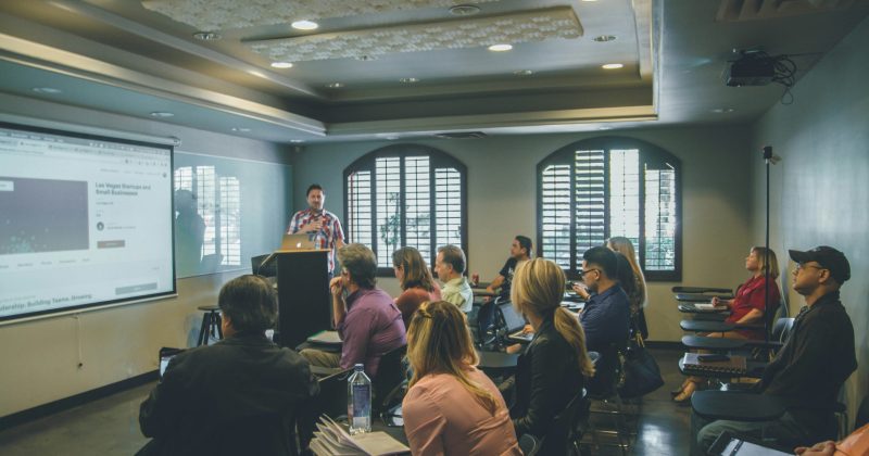 Speaker presenting to a group of professionals seated in a training room with a projected screen.