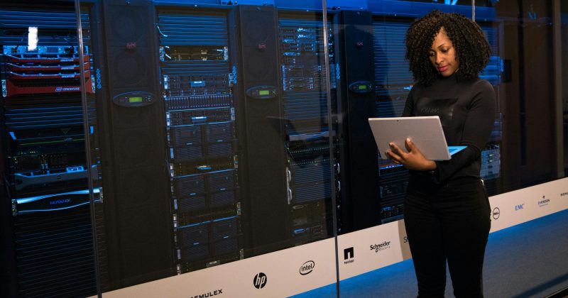 Woman using a laptop while standing in front of a server room filled with network equipment.