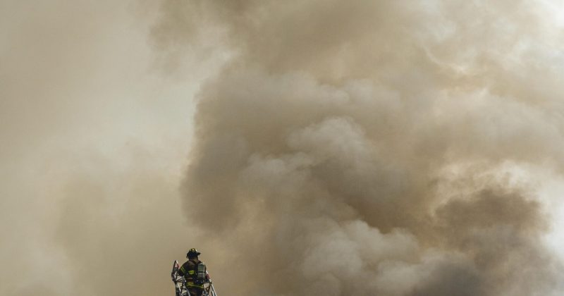 Firefighter atop an aerial ladder spraying water into thick smoke during a large-scale fire.